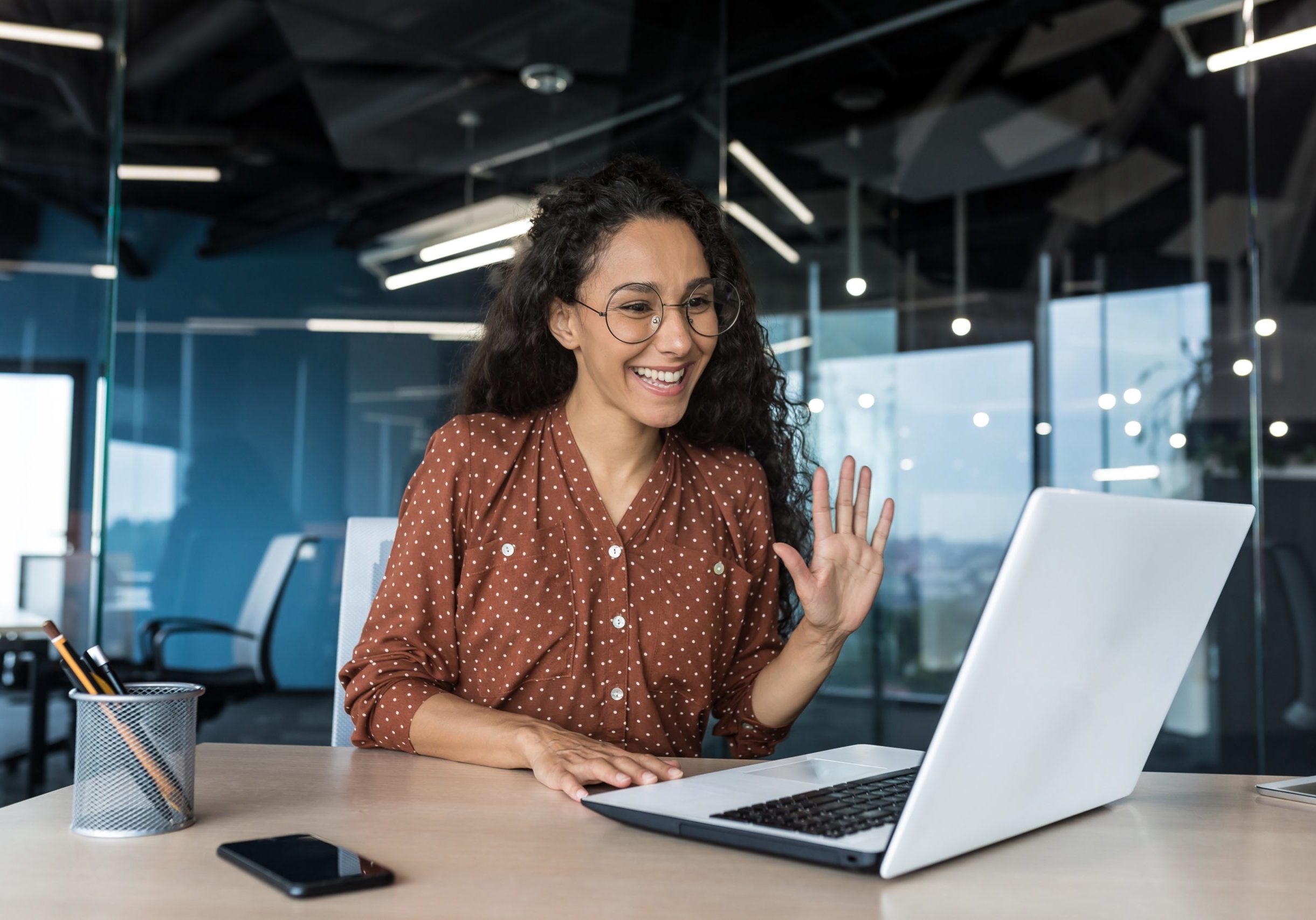 Successful hispanic woman working inside modern office building, businesswoman using laptop for video call smiling and waving, greeting gesture, online conference with colleagues